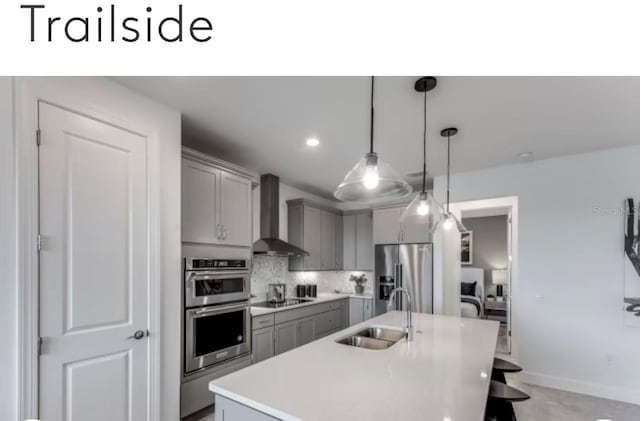 kitchen featuring gray cabinets, a sink, backsplash, appliances with stainless steel finishes, and wall chimney exhaust hood