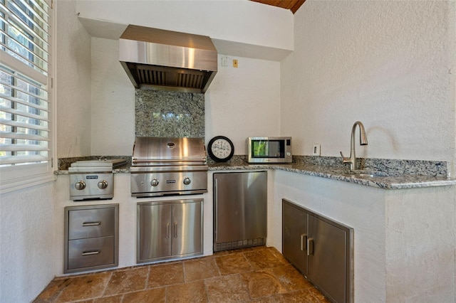 kitchen with stone countertops, a sink, wall chimney range hood, fridge, and stainless steel microwave