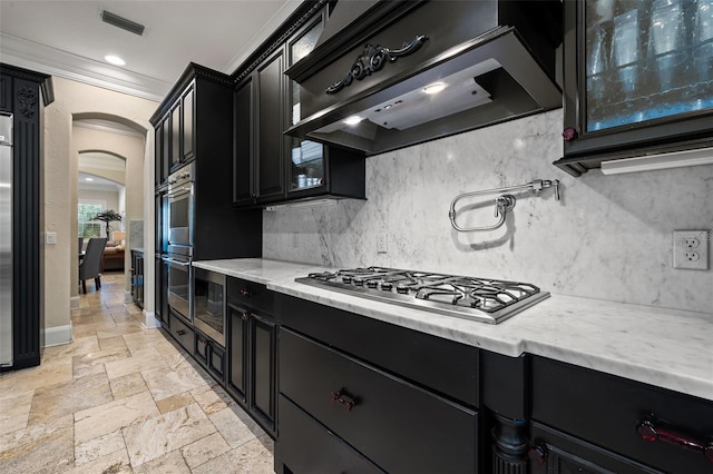 kitchen featuring stone tile floors, arched walkways, custom range hood, stainless steel appliances, and dark cabinetry