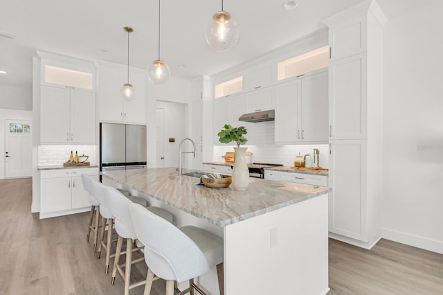 kitchen with under cabinet range hood, stainless steel appliances, a sink, a center island with sink, and glass insert cabinets