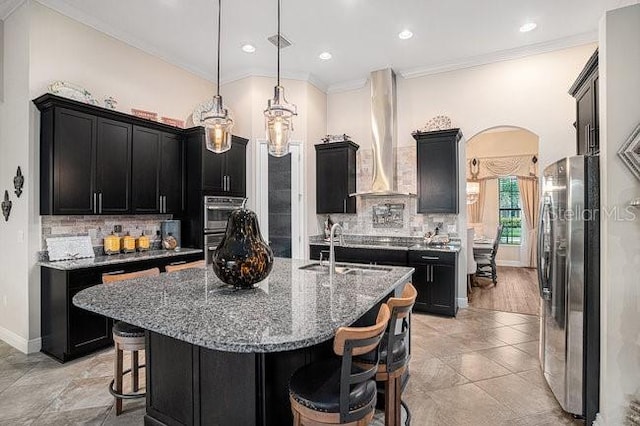 kitchen with arched walkways, dark cabinets, a kitchen breakfast bar, appliances with stainless steel finishes, and wall chimney exhaust hood