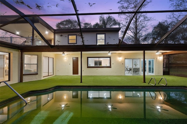 back of property at dusk featuring an outdoor pool, a lawn, glass enclosure, a patio area, and stucco siding