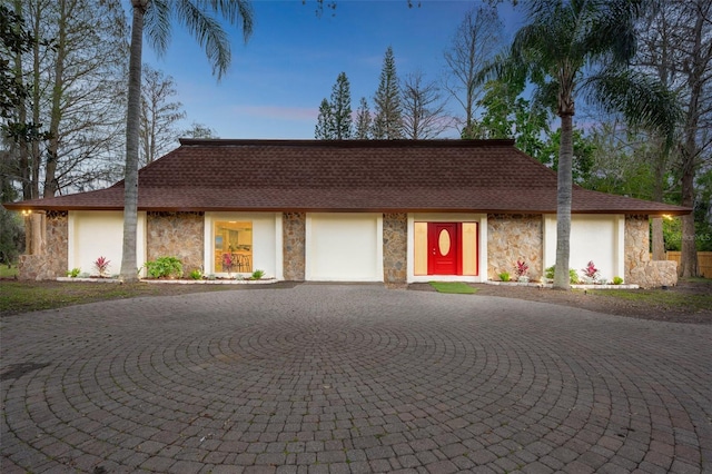 view of front of house with stone siding, roof with shingles, curved driveway, and stucco siding