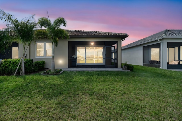 back of property at dusk featuring a sunroom, a tile roof, a lawn, and stucco siding