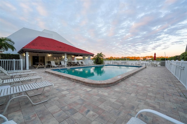 pool at dusk with a patio area, fence, and a community pool