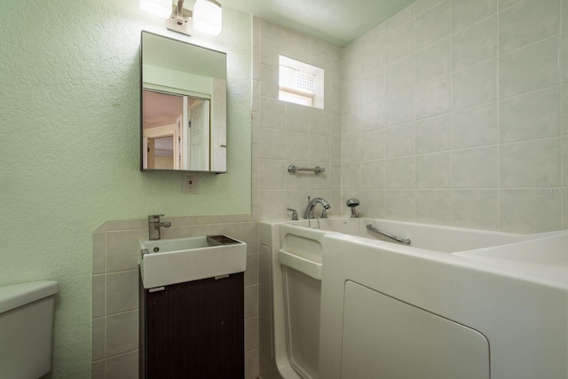 bathroom with a textured wall, toilet, vanity, tile walls, and a bath