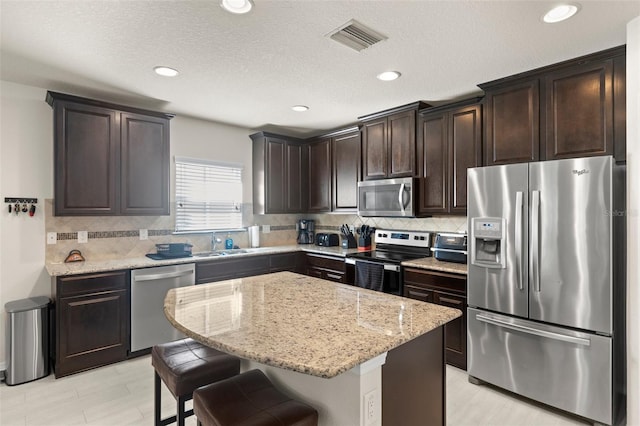 kitchen with dark brown cabinetry, visible vents, appliances with stainless steel finishes, light stone countertops, and tasteful backsplash