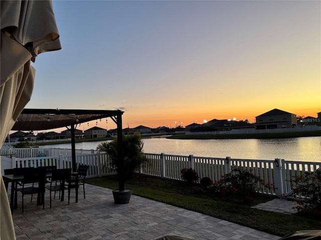 patio terrace at dusk featuring outdoor dining area, a water view, and a fenced backyard