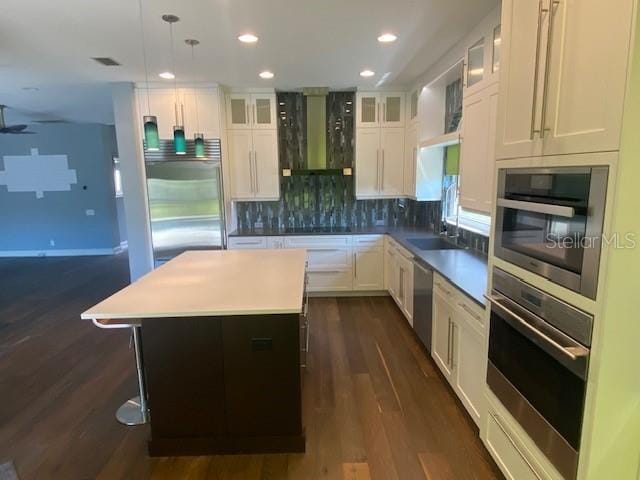 kitchen with tasteful backsplash, appliances with stainless steel finishes, white cabinetry, a kitchen island, and wall chimney exhaust hood