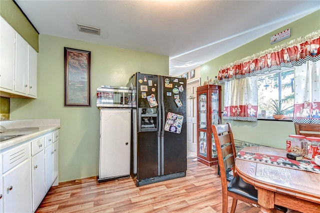 kitchen with light wood-type flooring, black fridge with ice dispenser, visible vents, and white cabinets