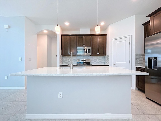 kitchen featuring arched walkways, dark brown cabinetry, stainless steel appliances, a sink, and a center island with sink