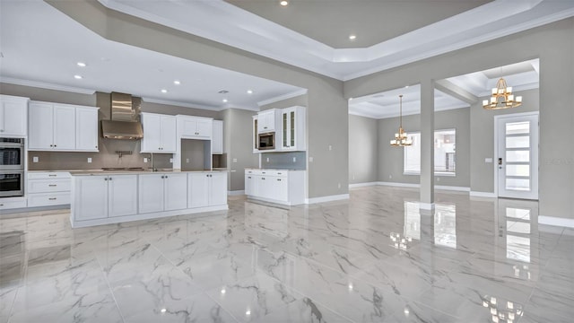 kitchen featuring a tray ceiling, a notable chandelier, stainless steel appliances, open floor plan, and wall chimney range hood