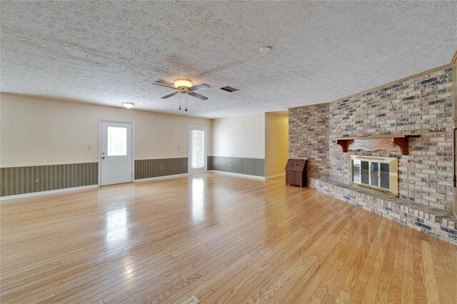 unfurnished living room featuring a textured ceiling, hardwood / wood-style flooring, a wainscoted wall, a ceiling fan, and a brick fireplace