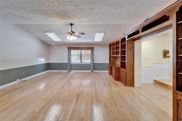 unfurnished living room featuring a textured ceiling, wainscoting, a ceiling fan, and light wood-style floors