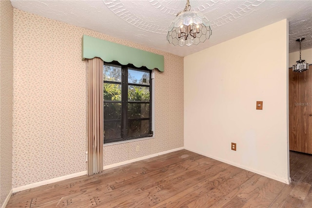 spare room with an inviting chandelier, a textured ceiling, wood finished floors, baseboards, and wallpapered walls