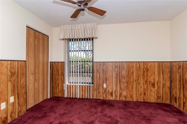 empty room with a textured ceiling, wooden walls, a wainscoted wall, carpet floors, and a ceiling fan