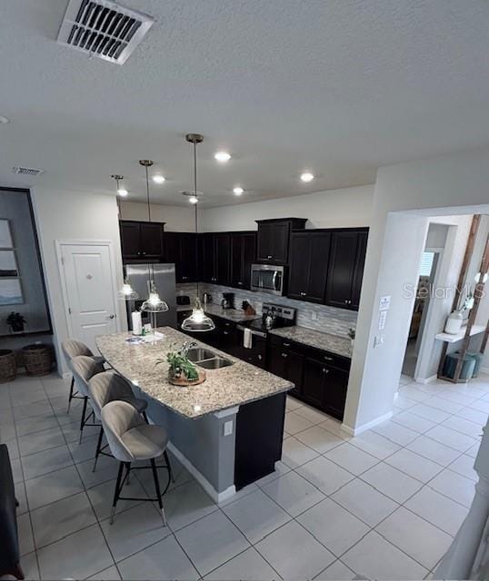 kitchen featuring visible vents, appliances with stainless steel finishes, dark cabinetry, backsplash, and a center island with sink