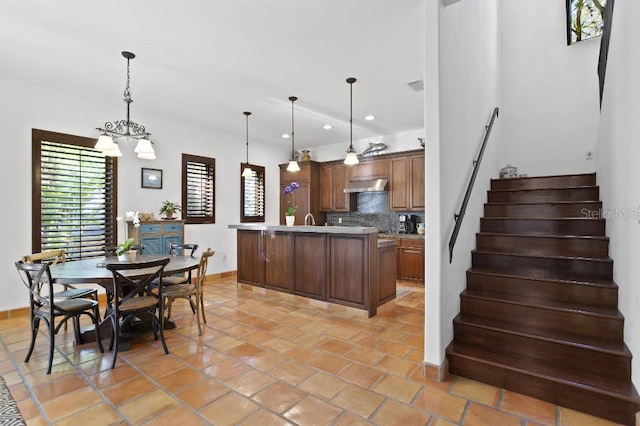 kitchen with tasteful backsplash, range hood, a kitchen island with sink, light countertops, and a notable chandelier