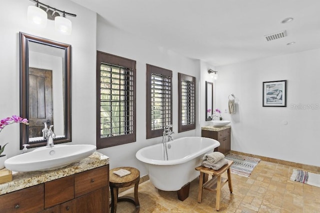 full bath featuring two vanities, visible vents, a freestanding bath, a sink, and baseboards