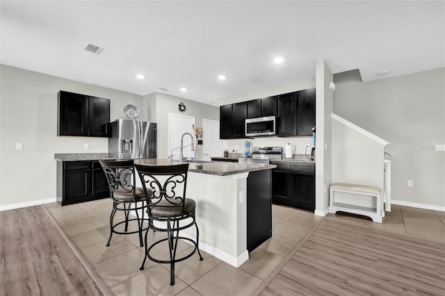 kitchen with an island with sink, appliances with stainless steel finishes, a kitchen breakfast bar, dark cabinets, and a sink