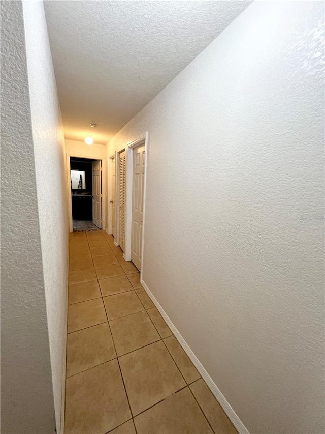 corridor featuring light tile patterned floors, a textured wall, a textured ceiling, and baseboards