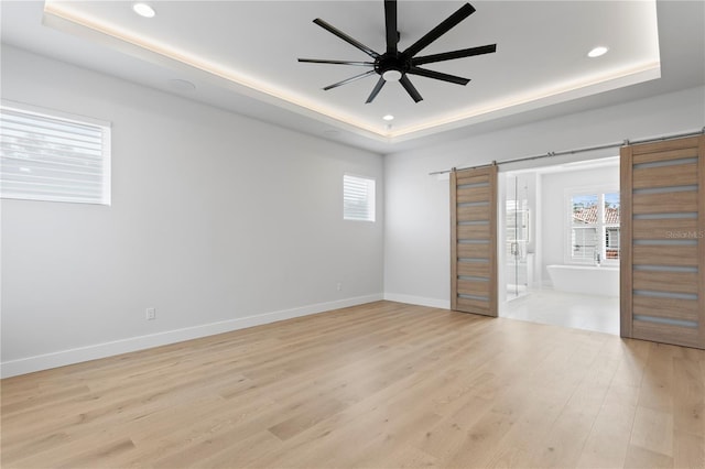 unfurnished bedroom featuring a tray ceiling, a barn door, multiple windows, and light wood-style flooring