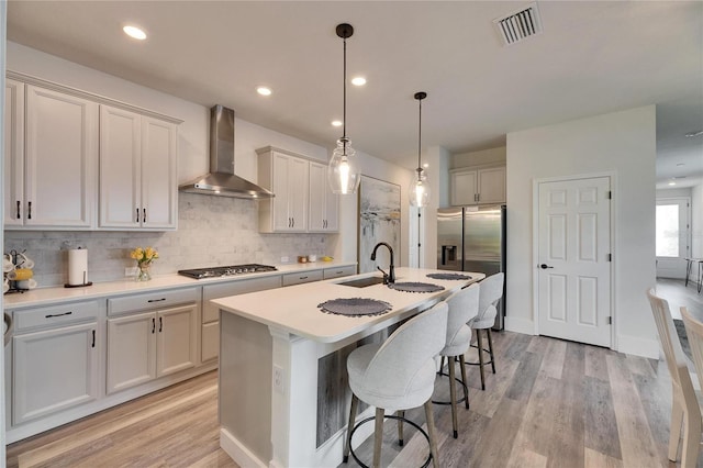 kitchen with visible vents, an island with sink, wall chimney exhaust hood, appliances with stainless steel finishes, and a sink