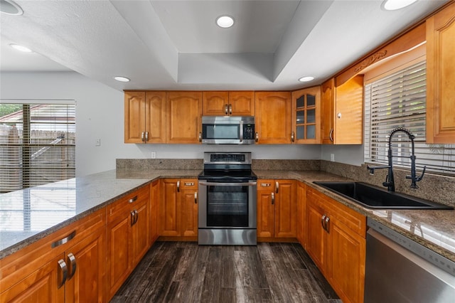 kitchen with light stone counters, stainless steel appliances, a sink, brown cabinets, and dark wood-style floors