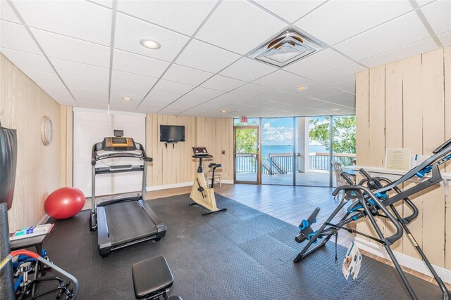 gym with expansive windows, wood walls, a drop ceiling, and baseboards