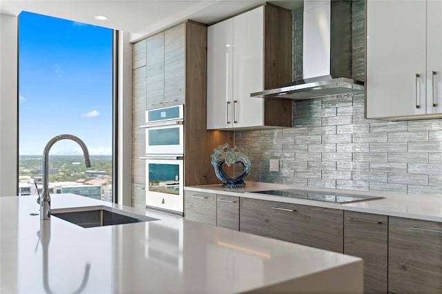 kitchen featuring black electric stovetop, white double oven, a sink, wall chimney range hood, and tasteful backsplash