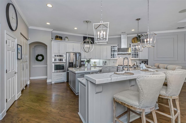 kitchen featuring a center island with sink, dark wood finished floors, wall chimney exhaust hood, stainless steel appliances, and a notable chandelier