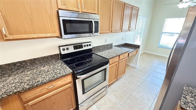 kitchen featuring baseboards, ceiling fan, appliances with stainless steel finishes, dark stone countertops, and light tile patterned floors