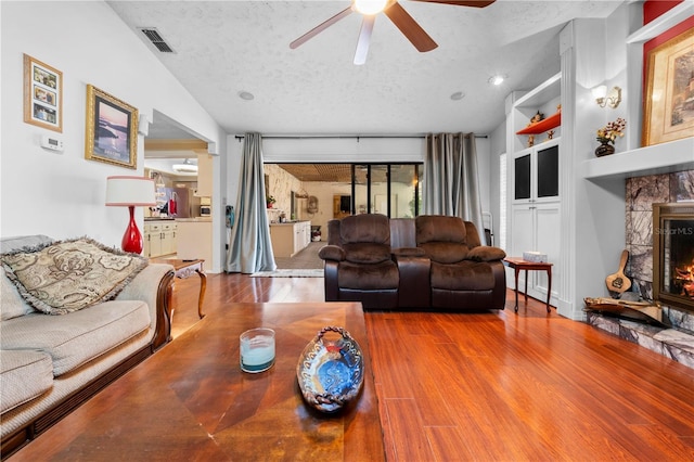 living area with a textured ceiling, a stone fireplace, wood finished floors, visible vents, and vaulted ceiling