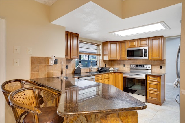 kitchen featuring dark stone counters, appliances with stainless steel finishes, a peninsula, a sink, and backsplash