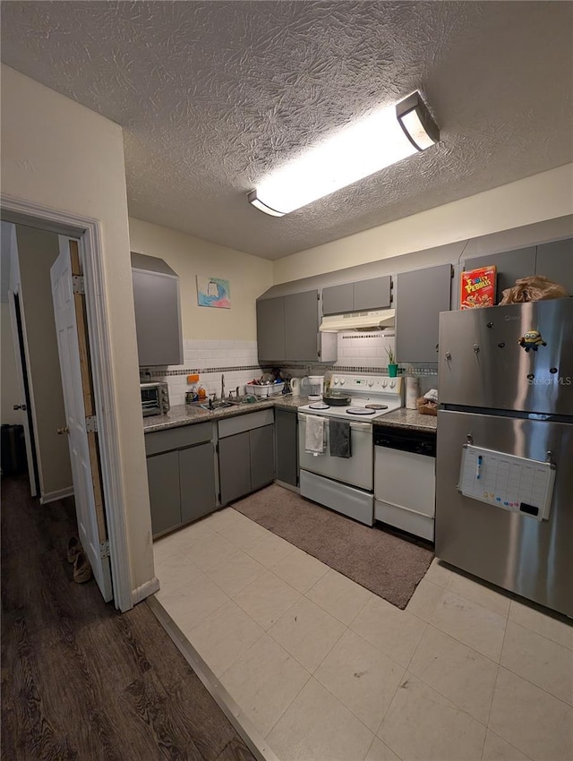 kitchen featuring under cabinet range hood, gray cabinetry, white appliances, a sink, and tasteful backsplash