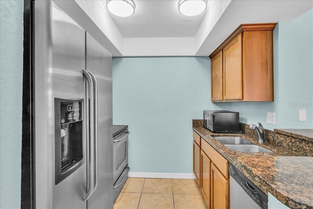 kitchen with appliances with stainless steel finishes, brown cabinetry, light tile patterned flooring, a sink, and baseboards