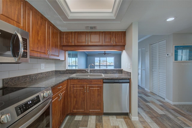 kitchen featuring a tray ceiling, crown molding, stainless steel appliances, brown cabinetry, and a sink