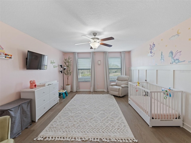 bedroom featuring ceiling fan, a textured ceiling, a wainscoted wall, wood finished floors, and a nursery area