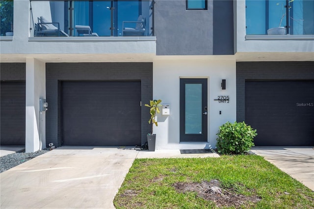 doorway to property featuring brick siding, stucco siding, concrete driveway, an attached garage, and a balcony