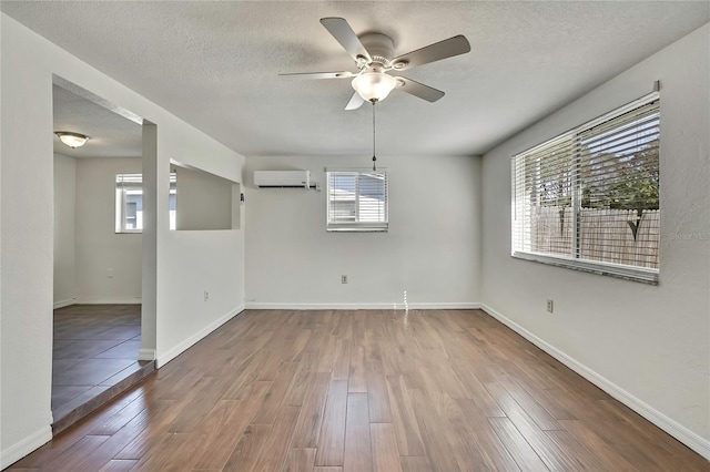 empty room with a textured ceiling, wood finished floors, a ceiling fan, baseboards, and an AC wall unit