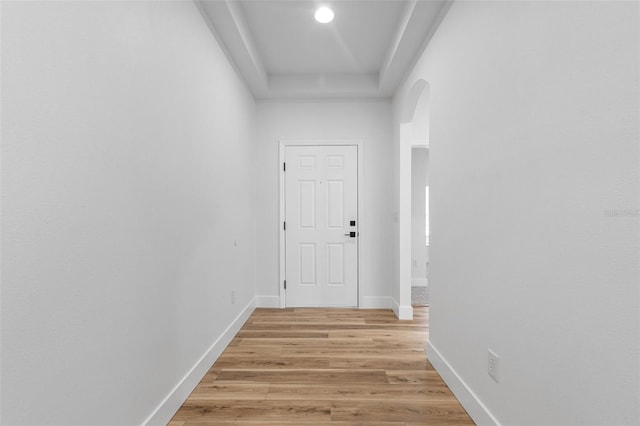 hall with arched walkways, a raised ceiling, baseboards, and light wood finished floors