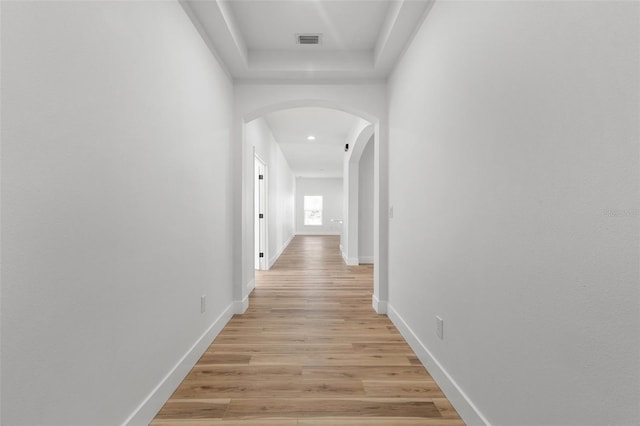 hall featuring light wood finished floors, baseboards, visible vents, arched walkways, and a tray ceiling