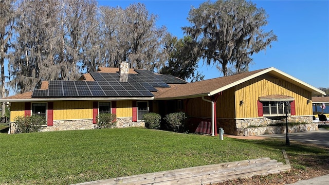 ranch-style house with solar panels, stone siding, a chimney, roof with shingles, and a front lawn