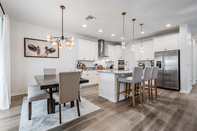 kitchen with stainless steel fridge, visible vents, an island with sink, wood finished floors, and wall chimney range hood