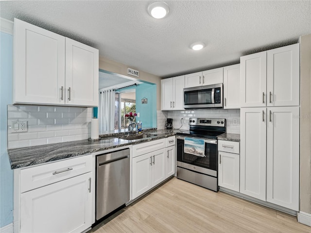 kitchen with stainless steel appliances, a sink, white cabinetry, light wood finished floors, and dark stone countertops