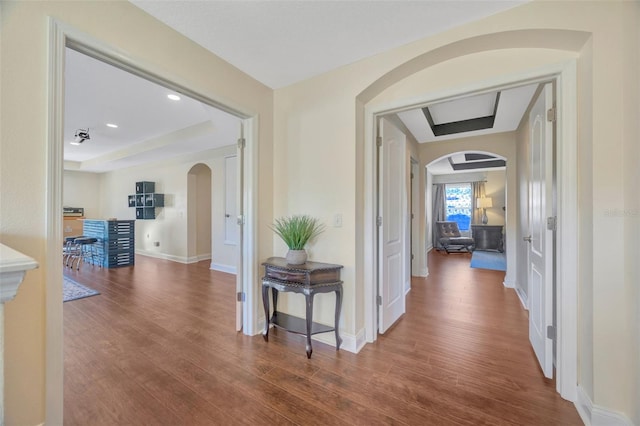 hallway with arched walkways, a tray ceiling, baseboards, and wood finished floors