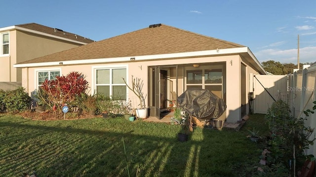 rear view of house featuring roof with shingles, a lawn, fence, and stucco siding