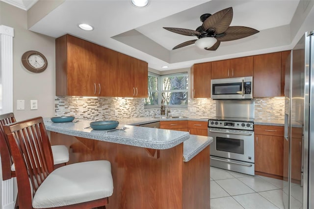 kitchen featuring a peninsula, a sink, appliances with stainless steel finishes, brown cabinets, and a tray ceiling