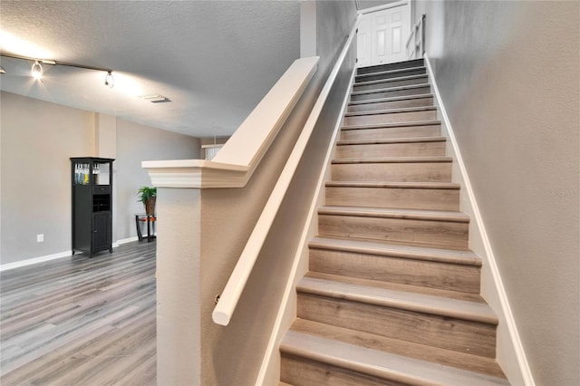 stairs featuring a textured ceiling, wood finished floors, rail lighting, and baseboards