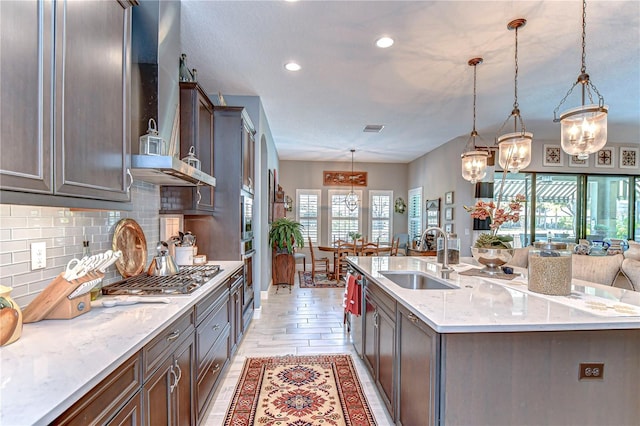 kitchen featuring light stone counters, a center island with sink, backsplash, a sink, and wall chimney exhaust hood